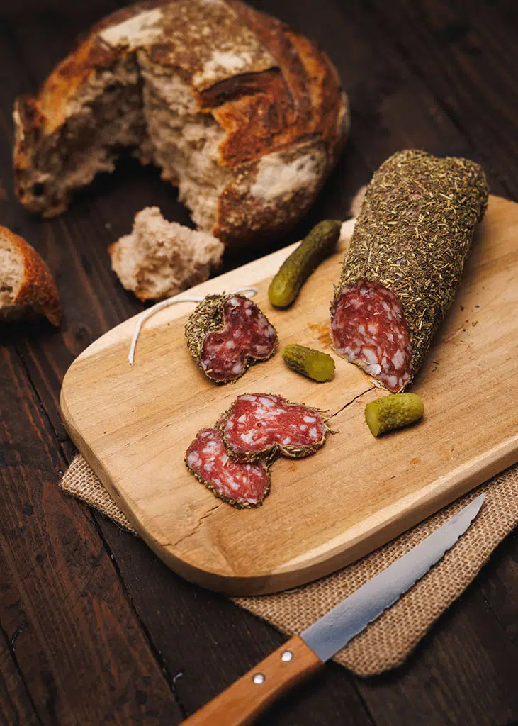 Product pack shot of sliced Provençal herb sausage on a wooden board — lifestyle photography of artisanal charcuterie against a dark wood background, Paris