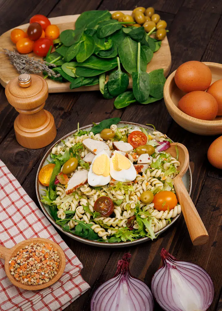 Food photography: mixed salad with pasta, chicken, hard-boiled eggs, cherry tomatoes, and olives — lifestyle styling on a wooden background, Paris