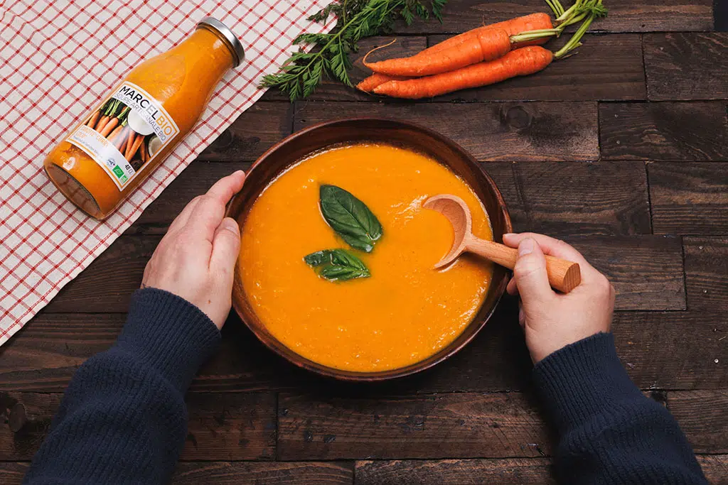 Product pack shot for Marcel Bio organic carrot soup — food photography featuring hands holding a terracotta bowl against a dark wooden background, Paris